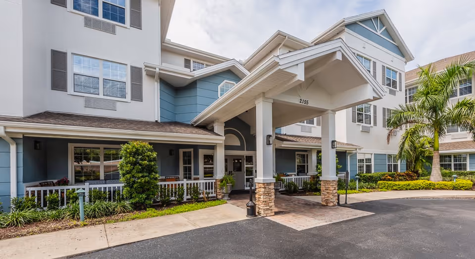Exterior front entrance of a senior living facility with a covered drop-off area supported by white columns with stone bases. The building is three stories tall with blue and white siding, multiple windows, and landscaping including bushes and palm trees.