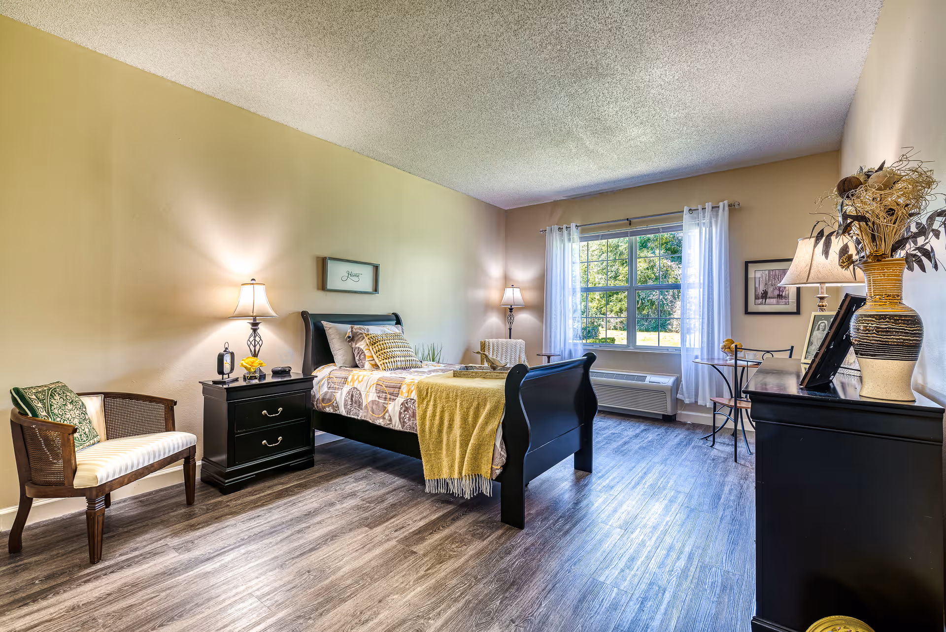 Well-lit furnished bedroom with a bed, nightstands, seating, dresser and a large window overlooking greenery.