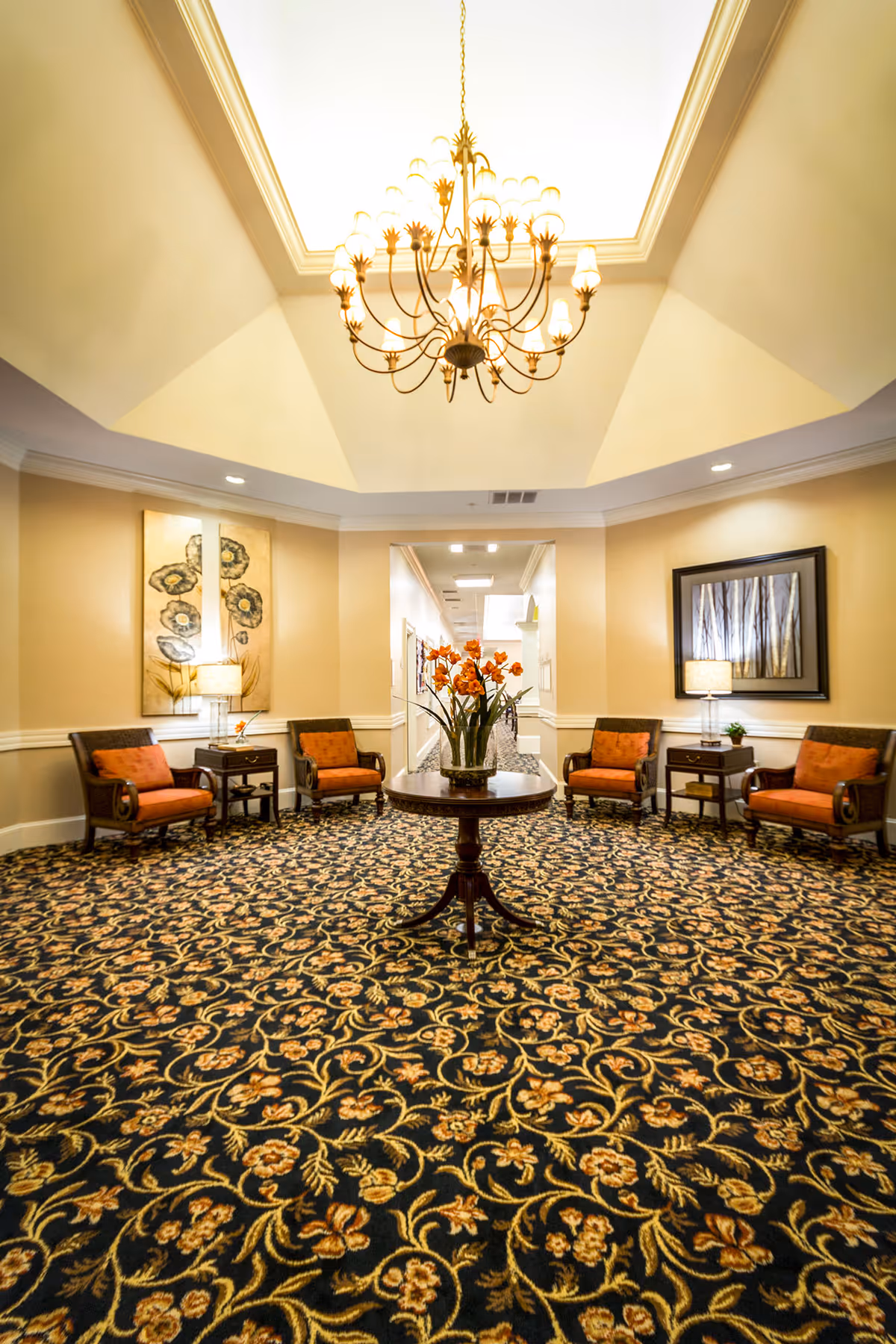 A spacious, well-lit sitting area with a patterned carpet, four armchairs with orange cushions arranged around two small side tables with lamps, a round wooden table with a vase of orange flowers in the center, and a large chandelier hanging from a recessed ceiling. The walls are decorated with framed artwork.