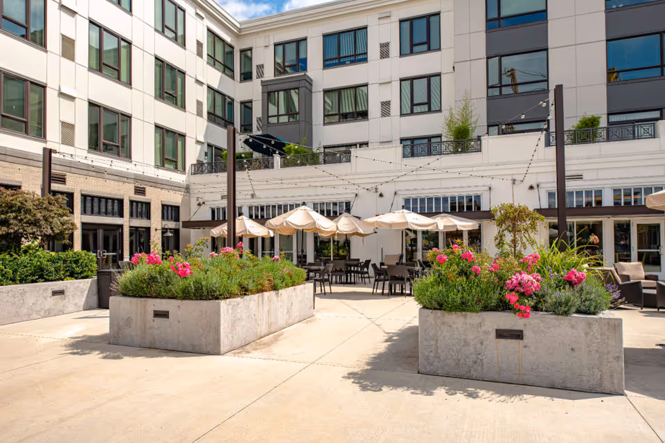 Outdoor courtyard area of a senior living facility with large concrete planters filled with green plants and pink flowers. Several tables with beige umbrellas are arranged for seating. The building surrounding the courtyard is multi-story with many windows, and string lights are hung above the seating area.