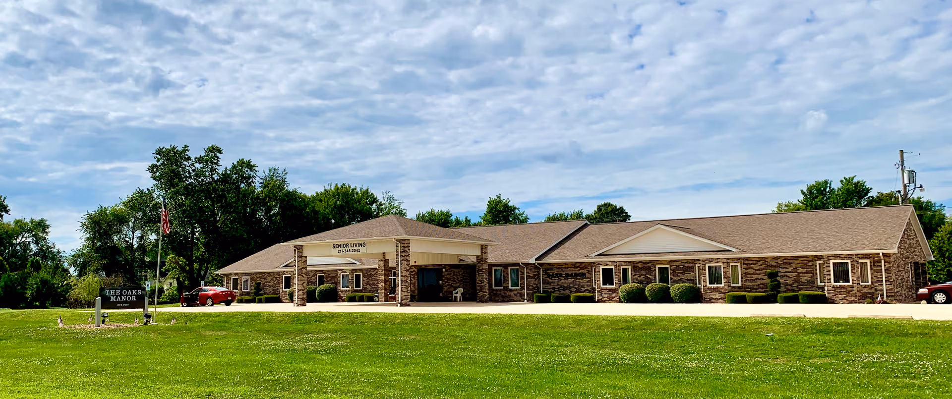 Single-story brick senior living building with a portico entrance, manicured lawn and a sign for The Oaks Manor.