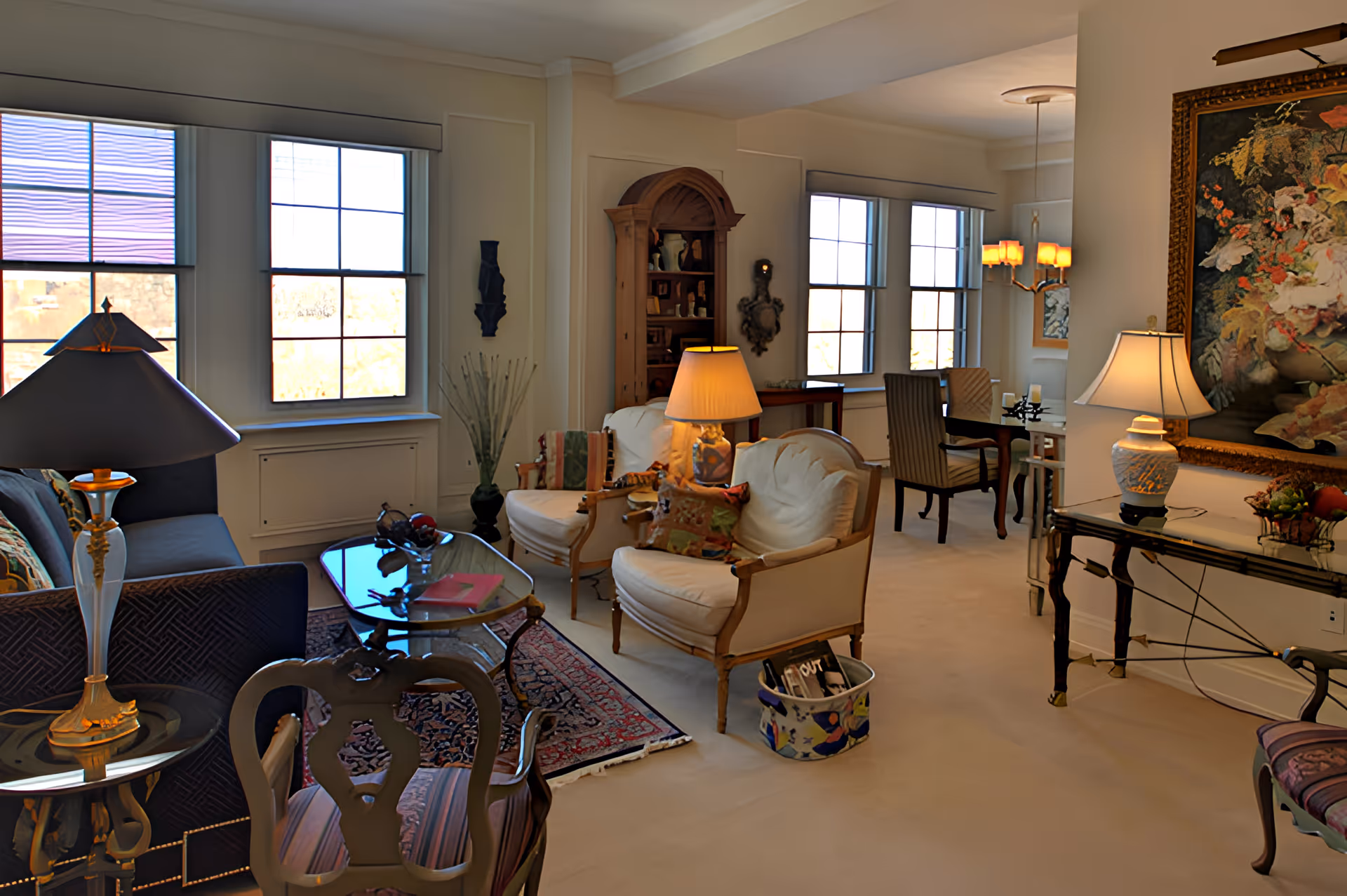 A cozy living room with multiple armchairs and a sofa arranged around a glass coffee table on a patterned rug. The room has large windows letting in natural light, several table lamps, a wooden cabinet with decorative items, and a large floral painting on the wall. There is also a dining area visible in the background with a table and chairs.