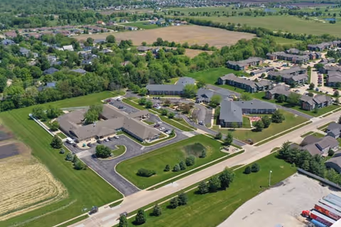 Aerial view of a senior living facility named Brookdale Urbana, showing multiple buildings surrounded by green lawns, trees, and parking areas, with residential neighborhoods and open fields in the background.