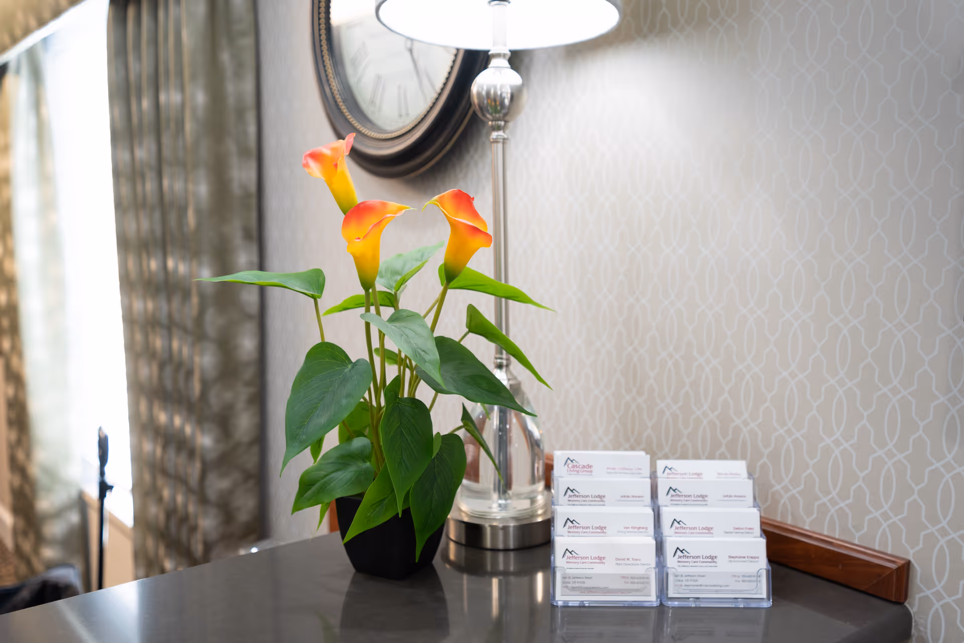 A close-up view of a countertop with a potted plant featuring orange and yellow flowers, a silver lamp, and a display of business cards for Jefferson Lodge Memory Care Community. A round wall clock and patterned wallpaper are visible in the background.