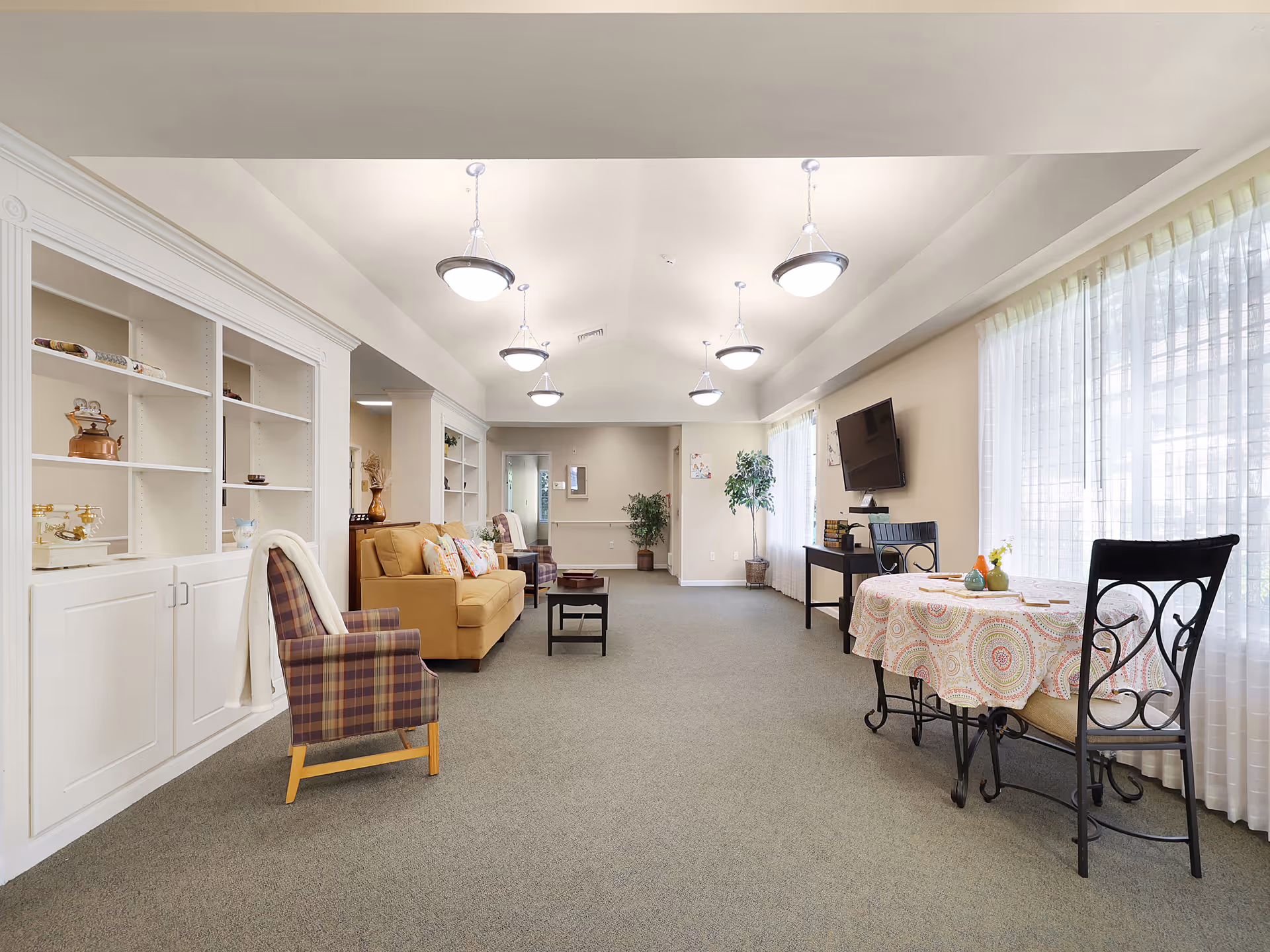 A bright and spacious common area in a senior living facility featuring a mustard yellow sofa with patterned pillows, a plaid armchair with a white throw, built-in white shelving units, a small round table with a colorful tablecloth and two chairs, a flat-screen TV mounted on the wall, and large windows with sheer white curtains allowing natural light to fill the room.