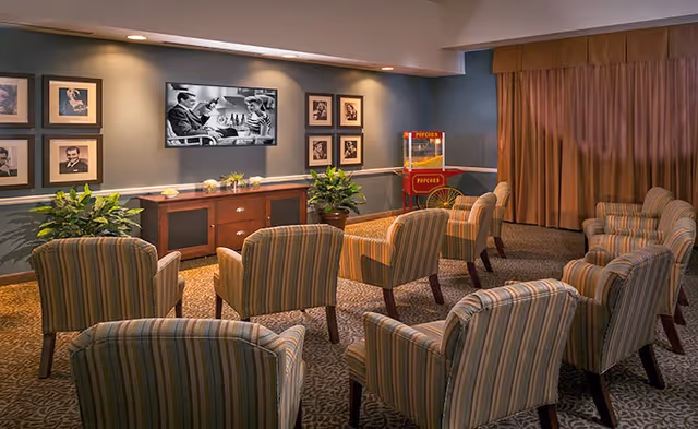 A cozy screening room with rows of striped armchairs facing a wall-mounted TV, framed photos, plants, and a popcorn machine.