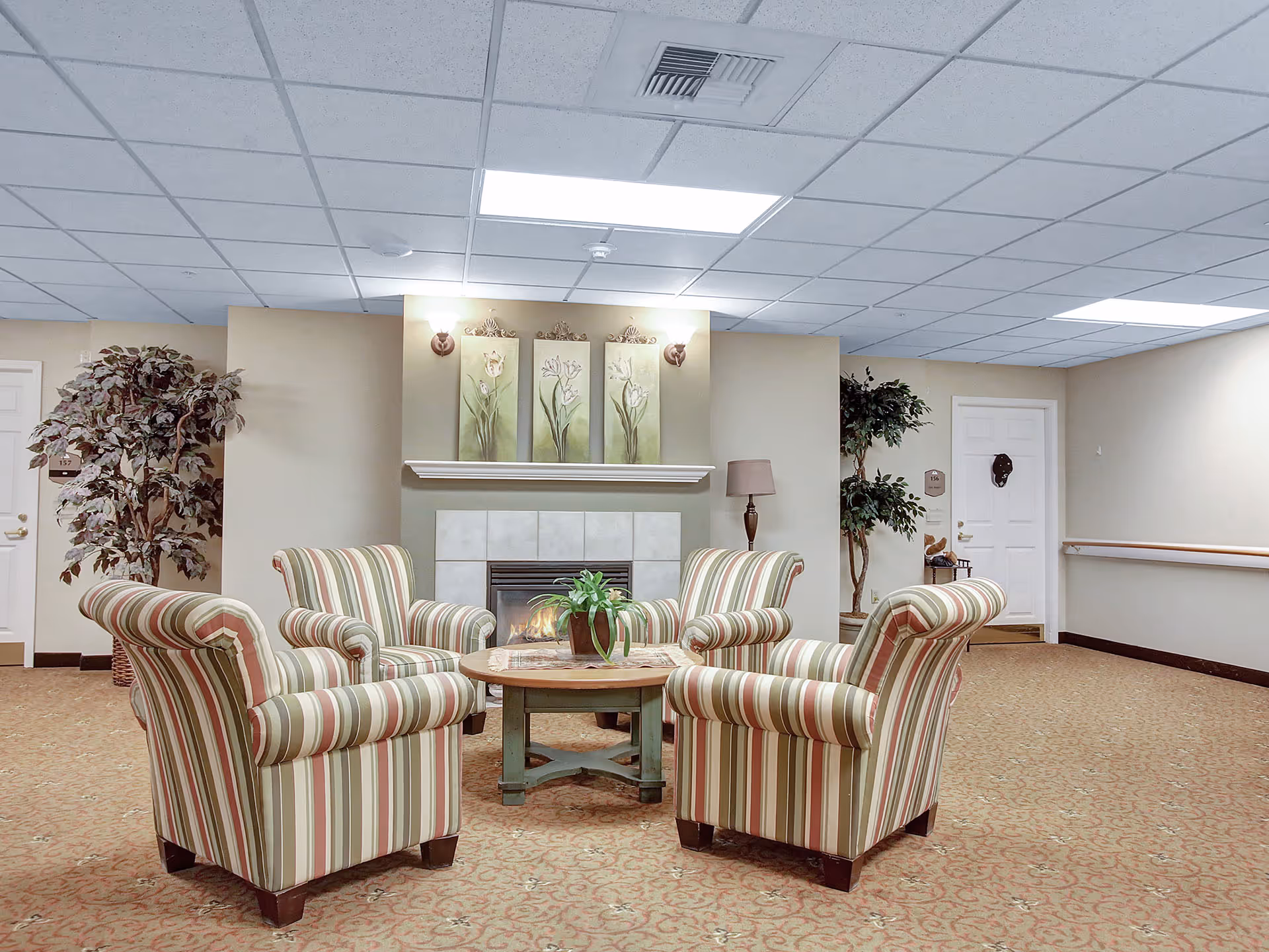 A cozy sitting area in a senior living facility featuring four striped armchairs arranged around a round wooden coffee table with a potted plant. Behind the chairs is a fireplace with three floral paintings above it, flanked by wall sconces. The room has beige walls, a patterned carpet, two potted plants, and white doors in the background.