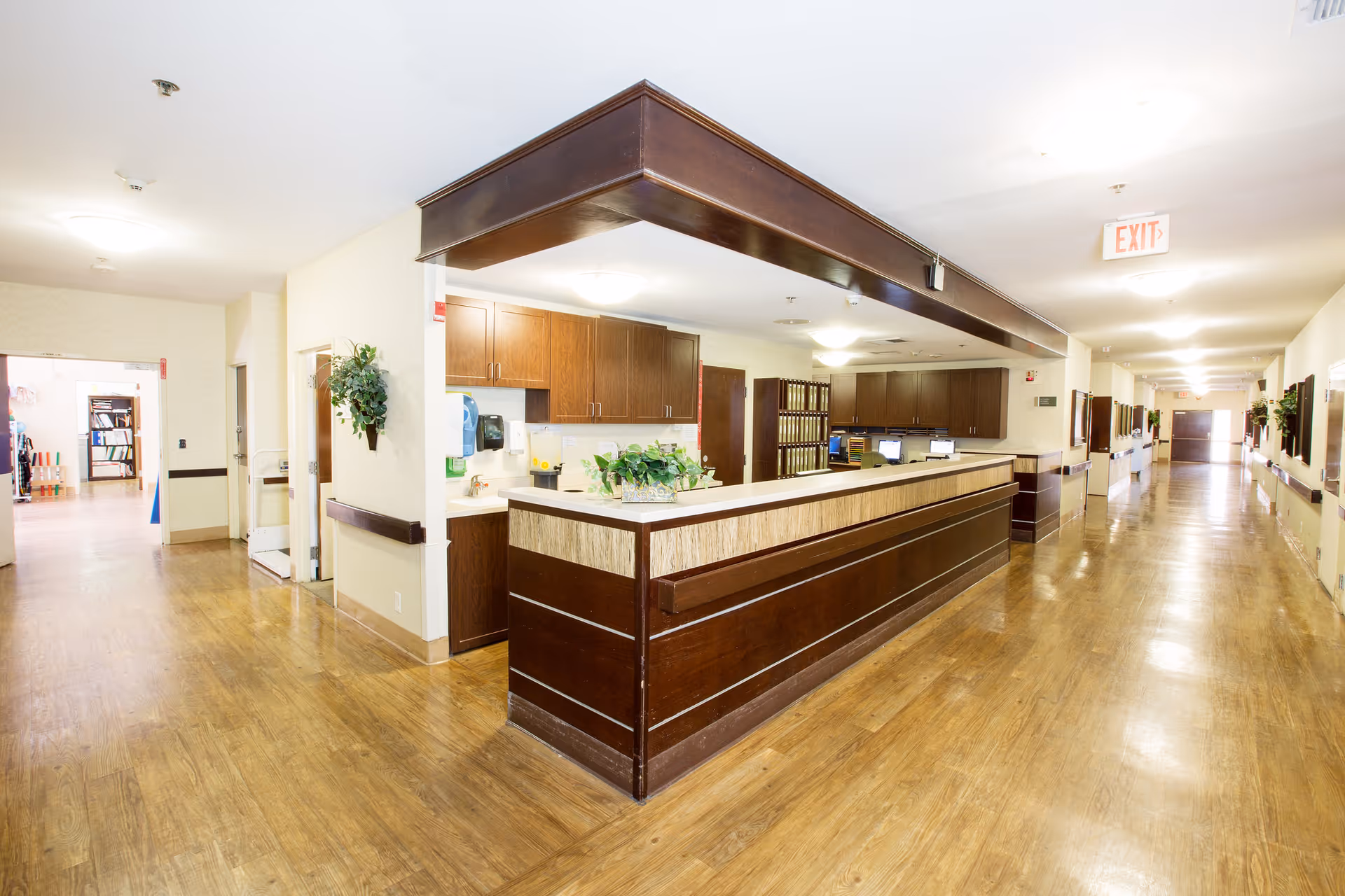 Interior view of a healthcare facility hallway with a long wooden reception desk in the center. The hallway has wooden flooring, cream-colored walls, and overhead lighting. There are plants mounted on the walls and computer monitors on the reception desk. Doors and exit signs are visible along the corridor.