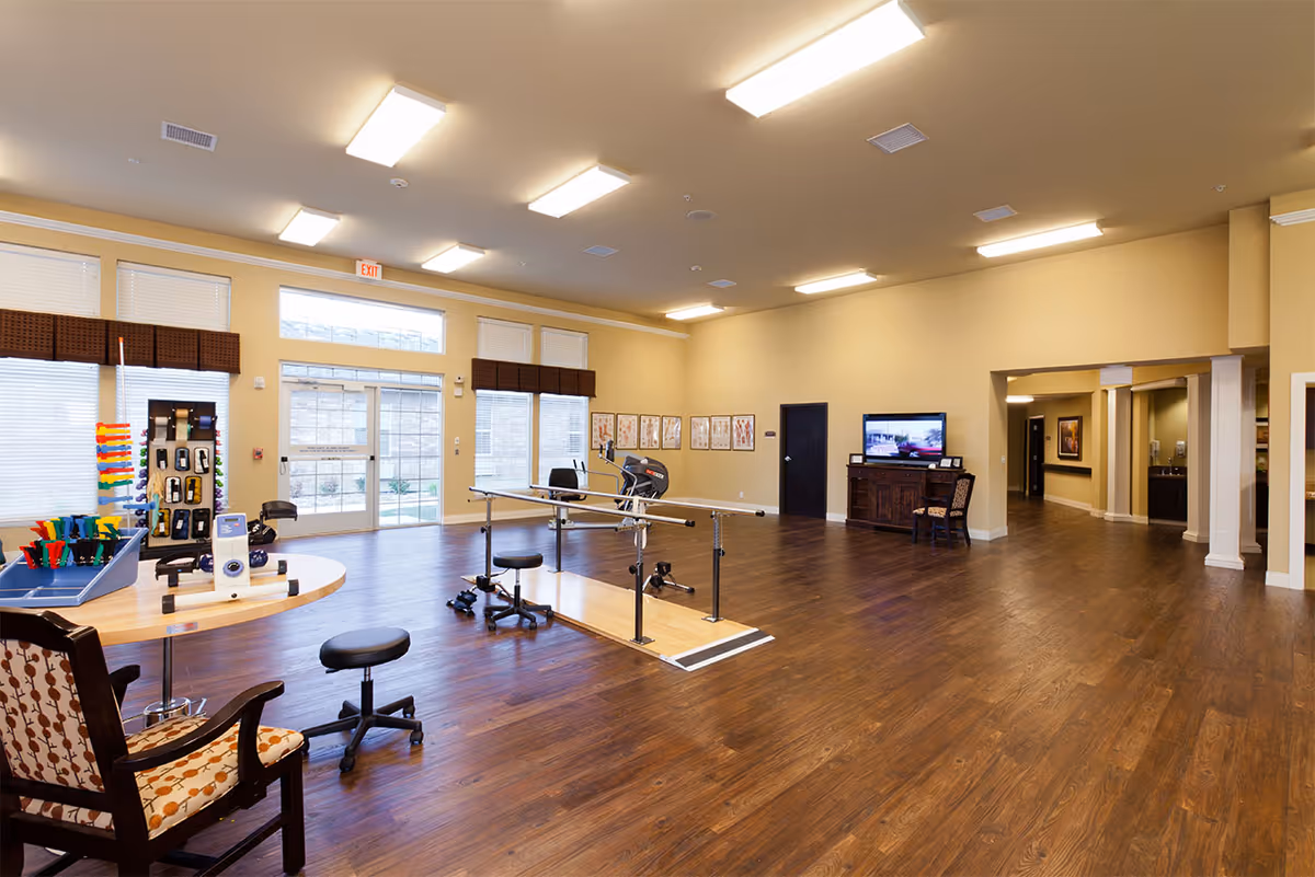 A spacious rehabilitation or therapy room with wooden flooring and beige walls. The room contains parallel bars for walking exercises, stools, a table with various therapy tools and equipment, a chair, and a TV on a wooden cabinet. Large windows and glass doors allow natural light to enter the room.