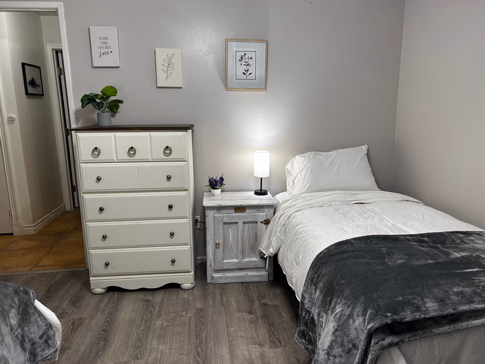A tidy single bedroom with a white dresser, a small nightstand and lamp, and a made bed with a gray throw.