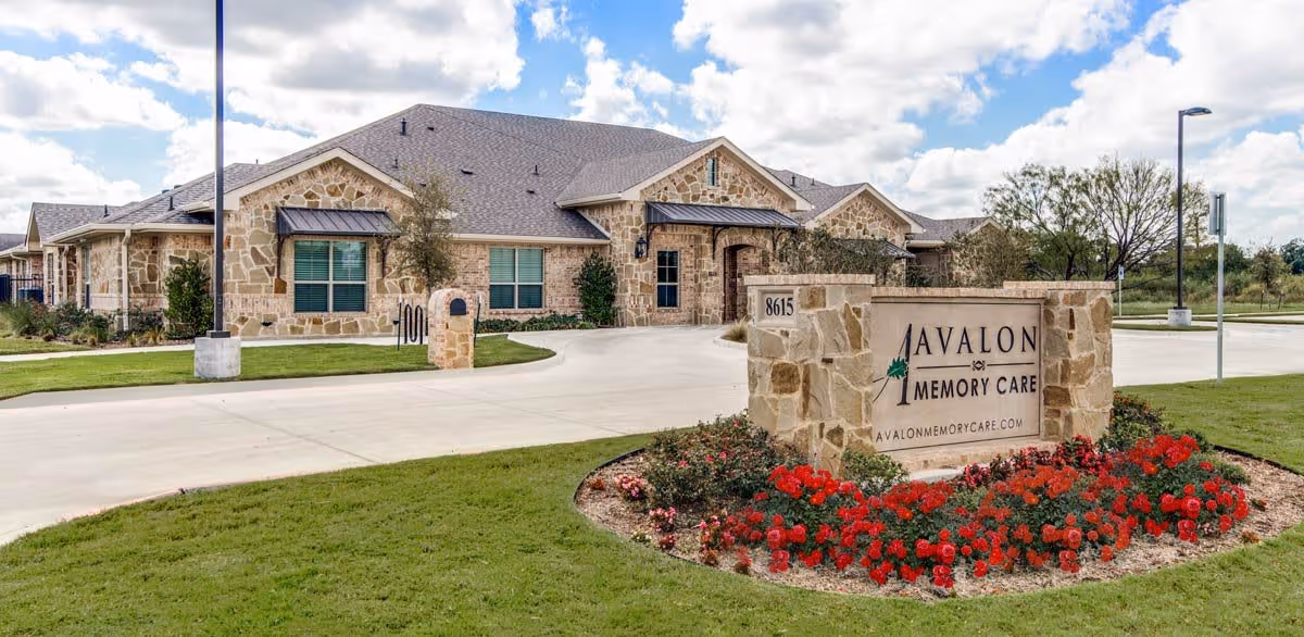 Exterior view of Avalon Memory Care facility showing a stone building with multiple windows and a driveway. In the foreground, there is a stone sign with the facility's name and website, surrounded by a flower bed with red flowers. The sky is partly cloudy.