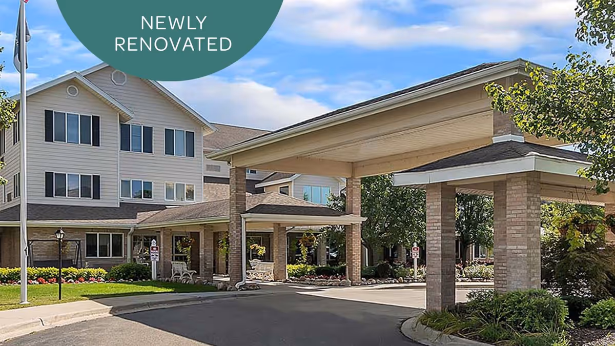 Exterior view of a newly renovated senior living facility building with a covered entrance driveway, surrounded by greenery and landscaping under a partly cloudy sky.