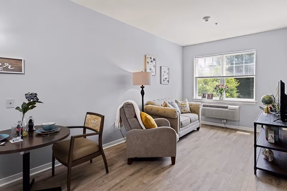 A bright living room with light gray walls and wood flooring. The room features a beige loveseat with decorative pillows, a gray armchair with a yellow pillow and a throw blanket, a floor lamp, and a window with blinds letting in natural light. A small round dining table with two chairs is set with plates, bowls, and a vase with flowers. A TV sits on a dark wooden stand with shelves holding books and decorative items.