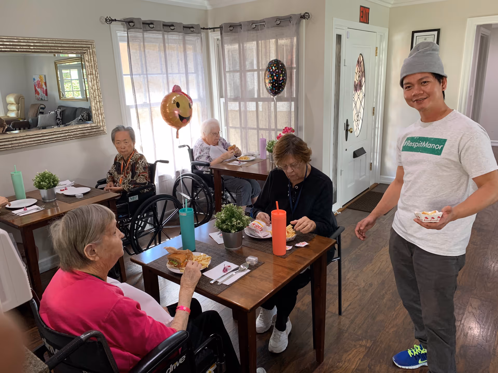 A group of elderly people, some in wheelchairs, are seated at tables in a dining area eating meals. A man wearing a gray beanie and a white t-shirt with the text #RespitManor stands nearby holding a small bowl of food. The room has wooden floors, large windows with sheer curtains, and a mirror on the wall. There are colorful balloons attached to some of the wheelchairs.