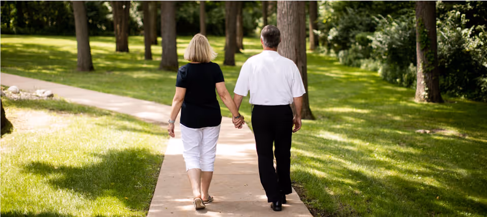 An elderly couple walking hand in hand on a paved path through a green park-like area with trees and grass on both sides.