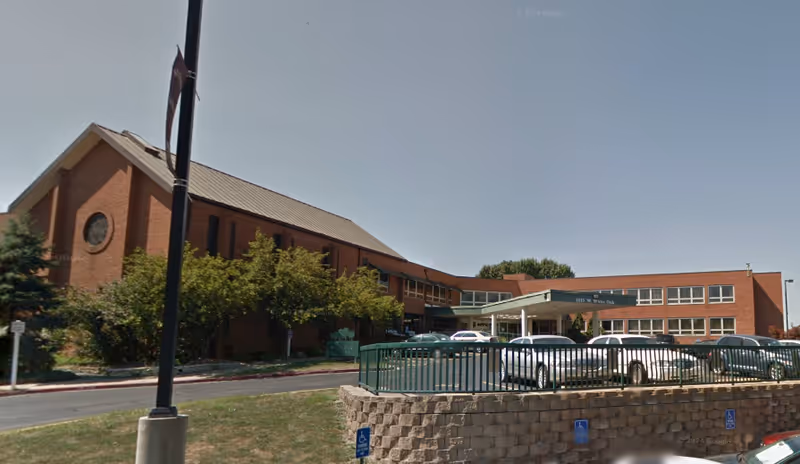 Brick senior living building with an entrance canopy and cars parked along the front.