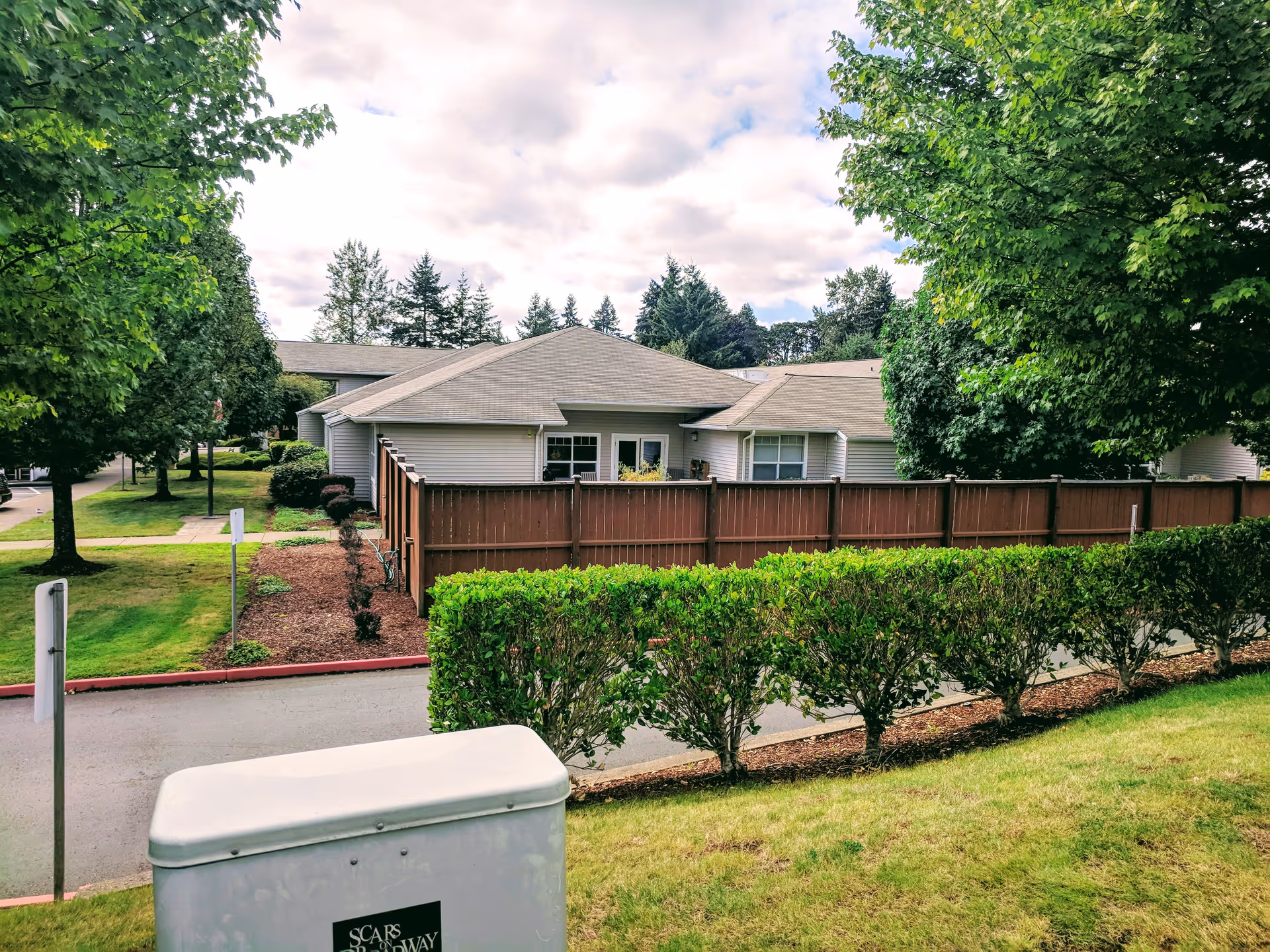 View of a single-story residential building with a brown wooden fence and green bushes in front, surrounded by trees and a grassy area under a partly cloudy sky.