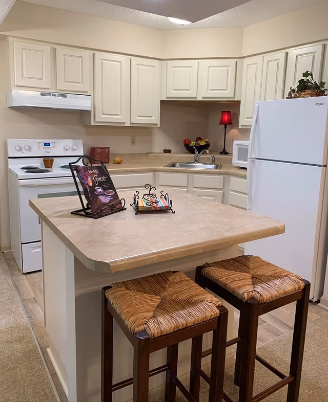 A clean and well-lit kitchen with white cabinets and appliances, including a stove, refrigerator, and microwave. There is a beige countertop island with two wooden stools that have woven seats. On the island, there is a decorative book stand holding a book and a small rack with colorful coasters. A small red lamp and a bowl of fruit are on the counter near the sink.