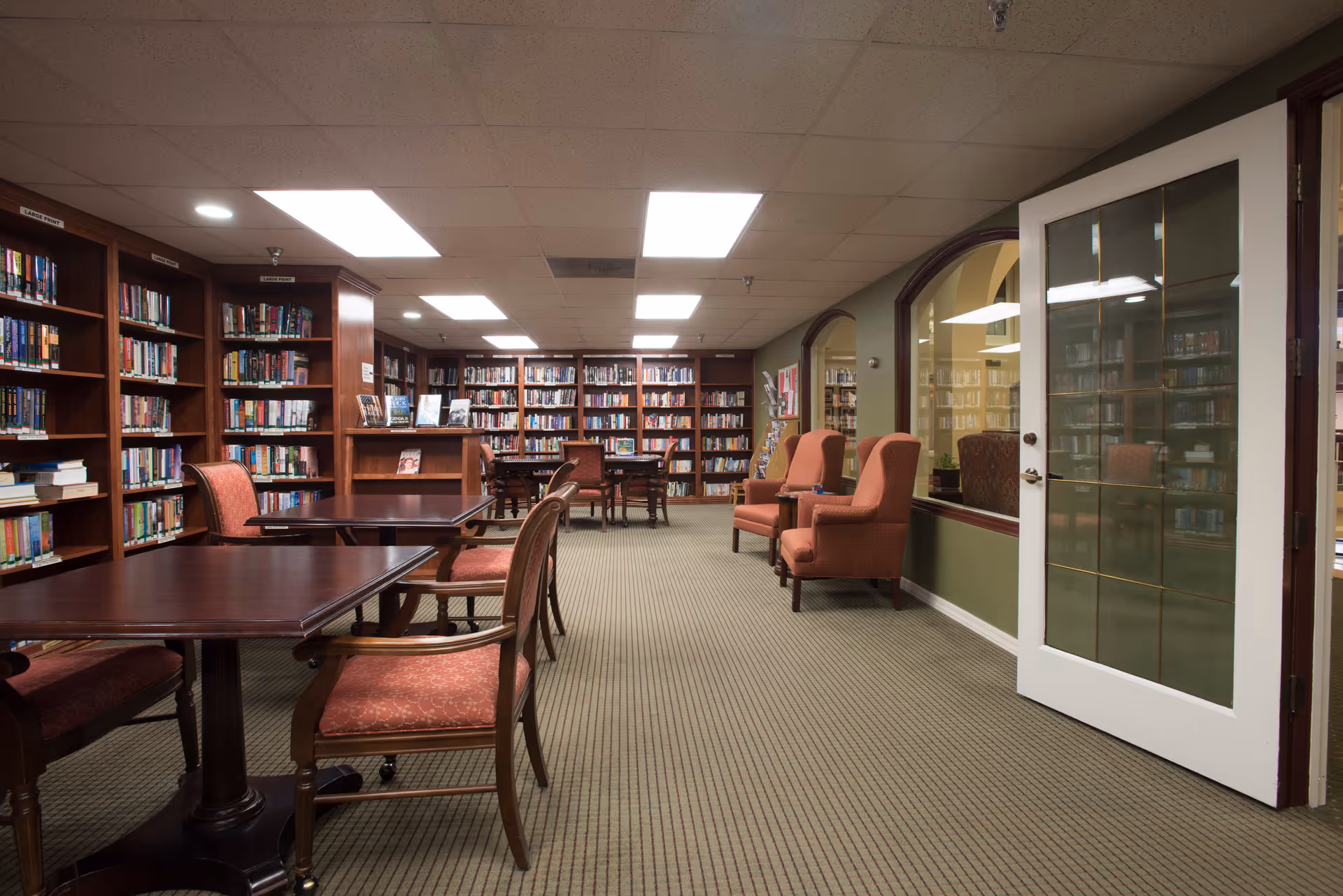 Interior view of a library room with wooden bookshelves filled with books along the walls, several wooden tables with upholstered chairs, and two upholstered armchairs near a wall with large windows and a white door with glass panels.