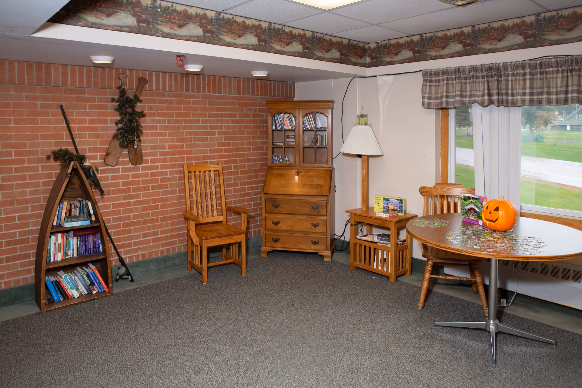 A cozy room with a brick wall, wooden furniture including a chair, a bookshelf shaped like a canoe filled with books, a wooden cabinet with glass doors, a small side table with a lamp, and a round table with a partially completed jigsaw puzzle and a carved pumpkin. A window with plaid curtains shows a view of green grass and a road outside. The ceiling has a decorative border with a nature scene.