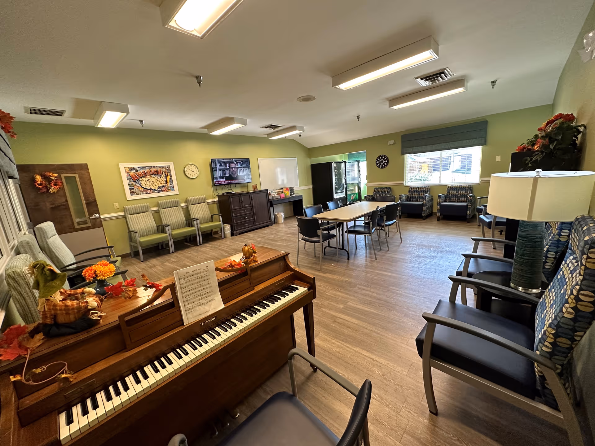 A spacious common room with green walls and wood flooring featuring a piano decorated with autumn-themed items, several chairs arranged along the walls, a table with chairs in the center, a television mounted on the wall, a dartboard, and windows letting in natural light.