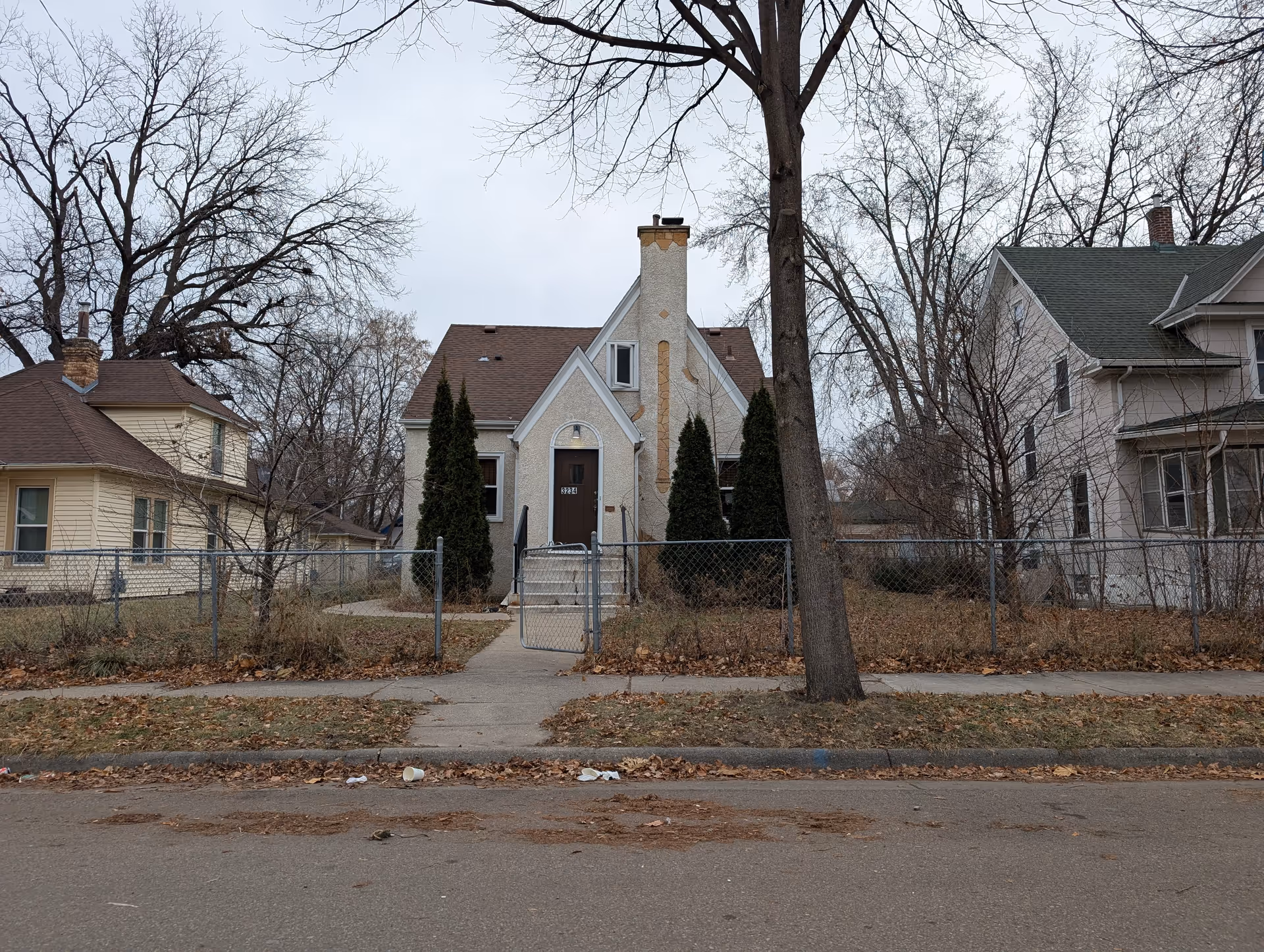 Front view of a small stucco house with a gated chain-link fence, leafless trees, and neighboring homes on a residential street.