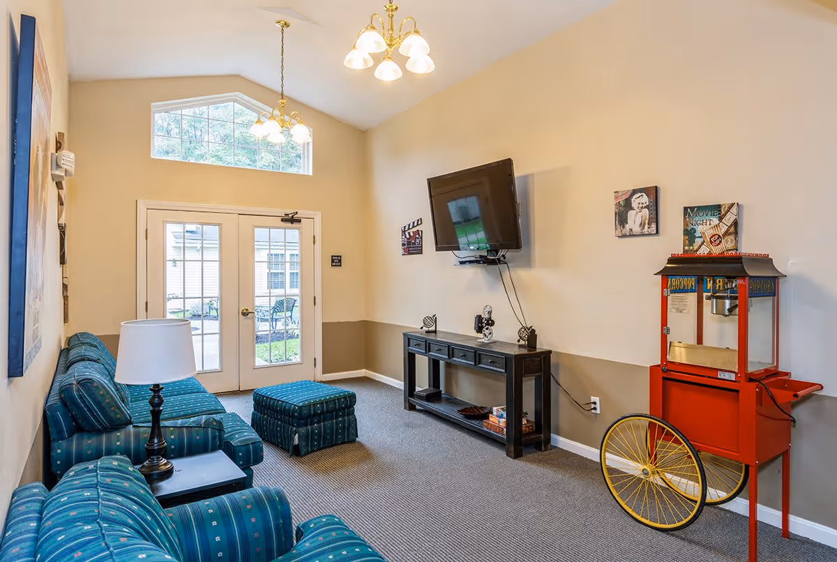 A cozy living room area with teal patterned sofas and an ottoman, a side table with a lamp, a wall-mounted flat-screen TV above a black console table, and a vintage-style red popcorn machine on wheels. The room has beige walls with a two-tone paint design, carpeted floor, and a set of glass double doors leading outside with a large window above them letting in natural light.