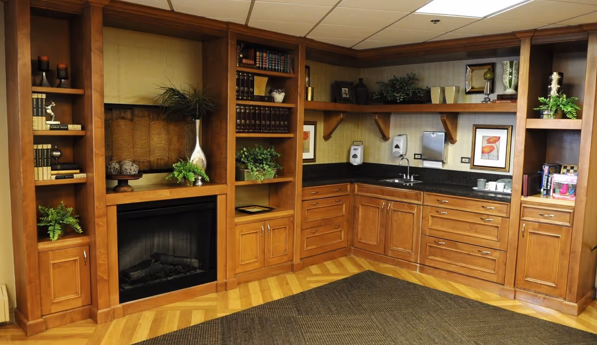 Cozy corner of a room featuring built-in wooden shelves and cabinets with decorative items, books, plants, and framed artwork. There is a small sink with soap dispensers and a paper towel holder on the countertop. A fireplace is integrated into the shelving unit. The floor has a wood pattern with a large dark rug.
