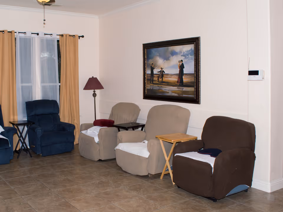 A sitting room with a row of recliner chairs, side tables, a floor lamp, and a framed painting on the wall.