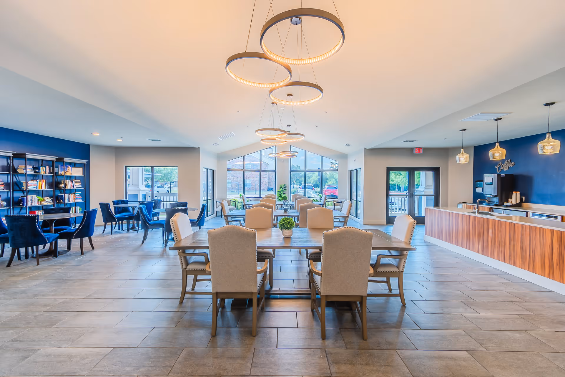 Bright and spacious common area with multiple seating arrangements including a large wooden table with beige upholstered chairs in the center, smaller round tables with blue chairs on the left, and a coffee station with pendant lights on the right. Large windows at the far end allow natural light to fill the room, and modern circular ceiling lights hang overhead.