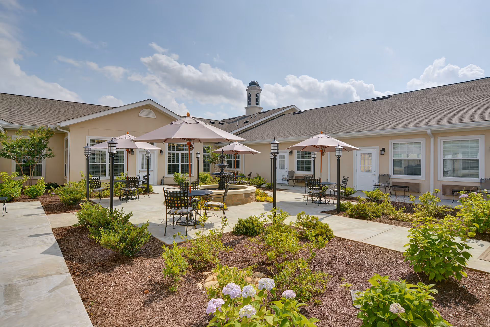Outdoor courtyard area of a senior living facility with several tables and chairs under umbrellas, surrounded by landscaped garden beds with shrubs and flowers, and beige buildings with white trim in the background under a partly cloudy sky.