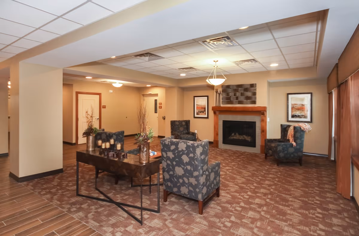 Spacious senior living common room with patterned armchairs, a central table, and a fireplace.
