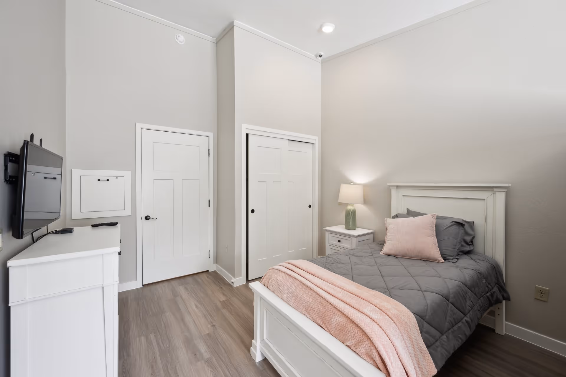 A neatly arranged bedroom with a white wooden bed frame, a gray quilted bedspread, and a pink throw blanket and pillow. There is a white nightstand with a green lamp beside the bed, a white dresser with a mounted flat-screen TV on the wall, and two white doors, one leading to a closet. The room has light gray walls and wood flooring.