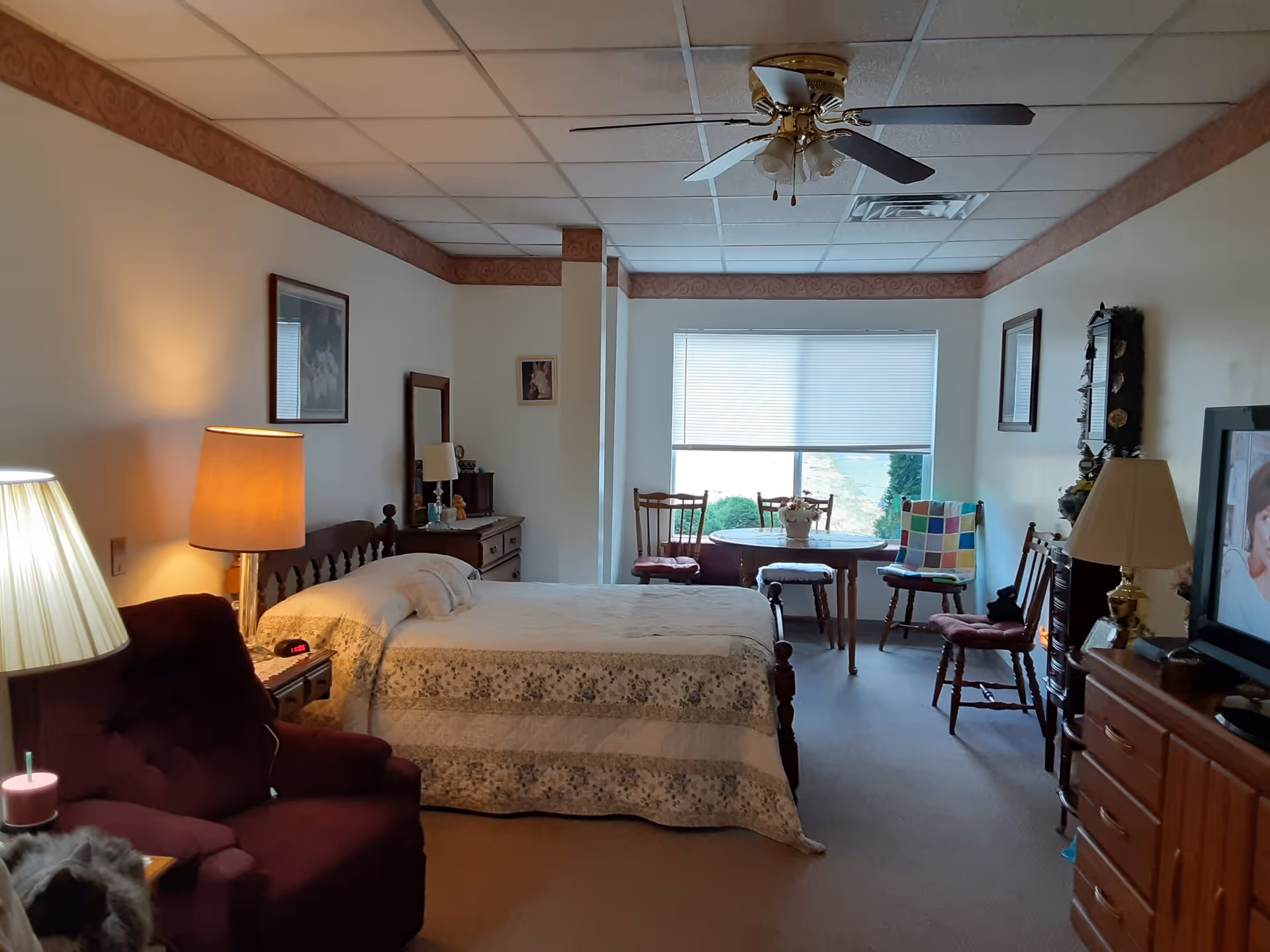 A cozy bedroom in a senior living facility with a bed covered in a floral quilt, a burgundy armchair with a side table and lamp, a dresser with a mirror, a round table with chairs near a window, and a television on a wooden cabinet. The room has soft lighting and a ceiling fan.