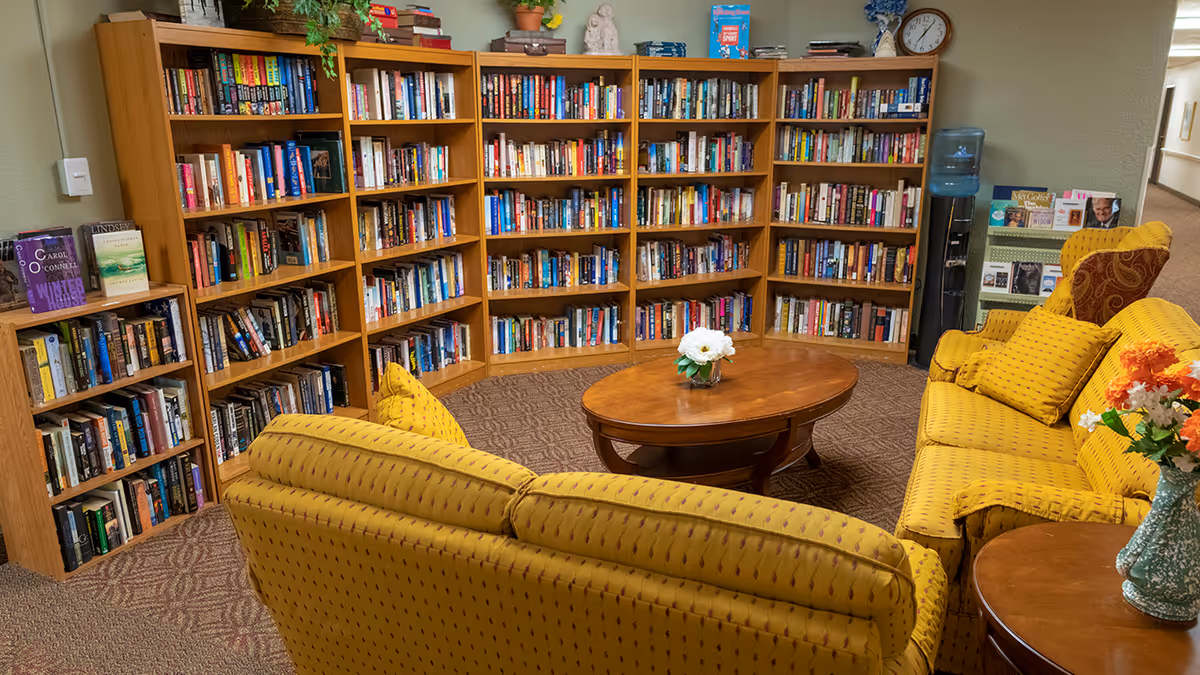 Cozy common library with bookshelves along the walls and yellow upholstered sofas arranged around a wooden coffee table.