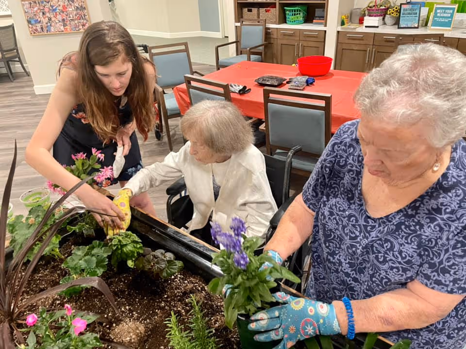 Three women engaged in indoor gardening activities around a raised planter filled with soil and various plants. One younger woman is assisting two elderly women, one in a wheelchair, as they plant flowers. The room has wooden flooring, chairs, and a table covered with a red cloth in the background.