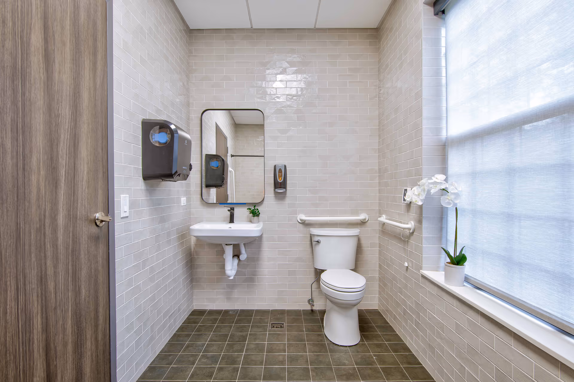 A clean, modern bathroom with beige tiled walls and dark tiled floor. It features a white toilet with grab bars on both sides, a white wall-mounted sink with a small green plant on it, a rectangular mirror above the sink, a soap dispenser, a paper towel dispenser, and a window with a sheer white curtain and a white orchid plant on the windowsill.