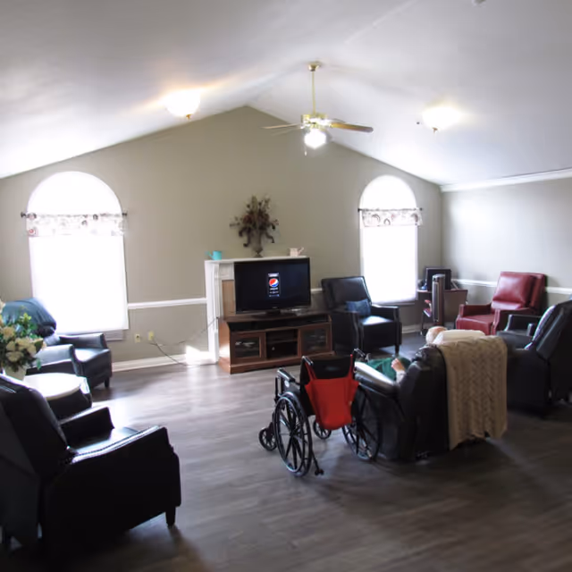A spacious living room with several armchairs arranged around a television on a wooden stand. There is a wheelchair with a red seat in the center of the room. The room has two large arched windows with valances, a ceiling fan with lights, and a decorative floral arrangement above the TV. The floor is wood, and the walls are painted light gray with white trim.