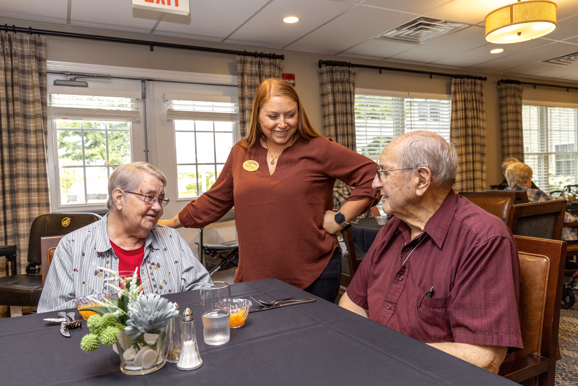 A staff member in a maroon shirt smiles and interacts with two elderly residents seated at a dining table with a black tablecloth. The table has a small floral centerpiece, salt and pepper shakers, and glasses of water. The room has large windows with plaid curtains and a warm, inviting atmosphere.
