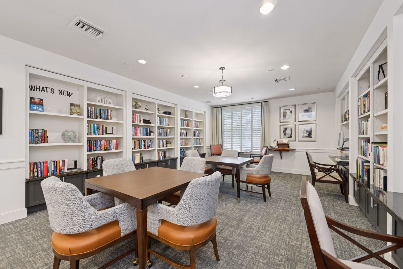 A bright and spacious room with multiple tables and chairs arranged for reading or group activities. The walls are lined with built-in bookshelves filled with books and decorative items. A window with white shutters and beige curtains lets in natural light. The room has a neutral color palette with gray carpeting and white walls, creating a calm and inviting atmosphere.