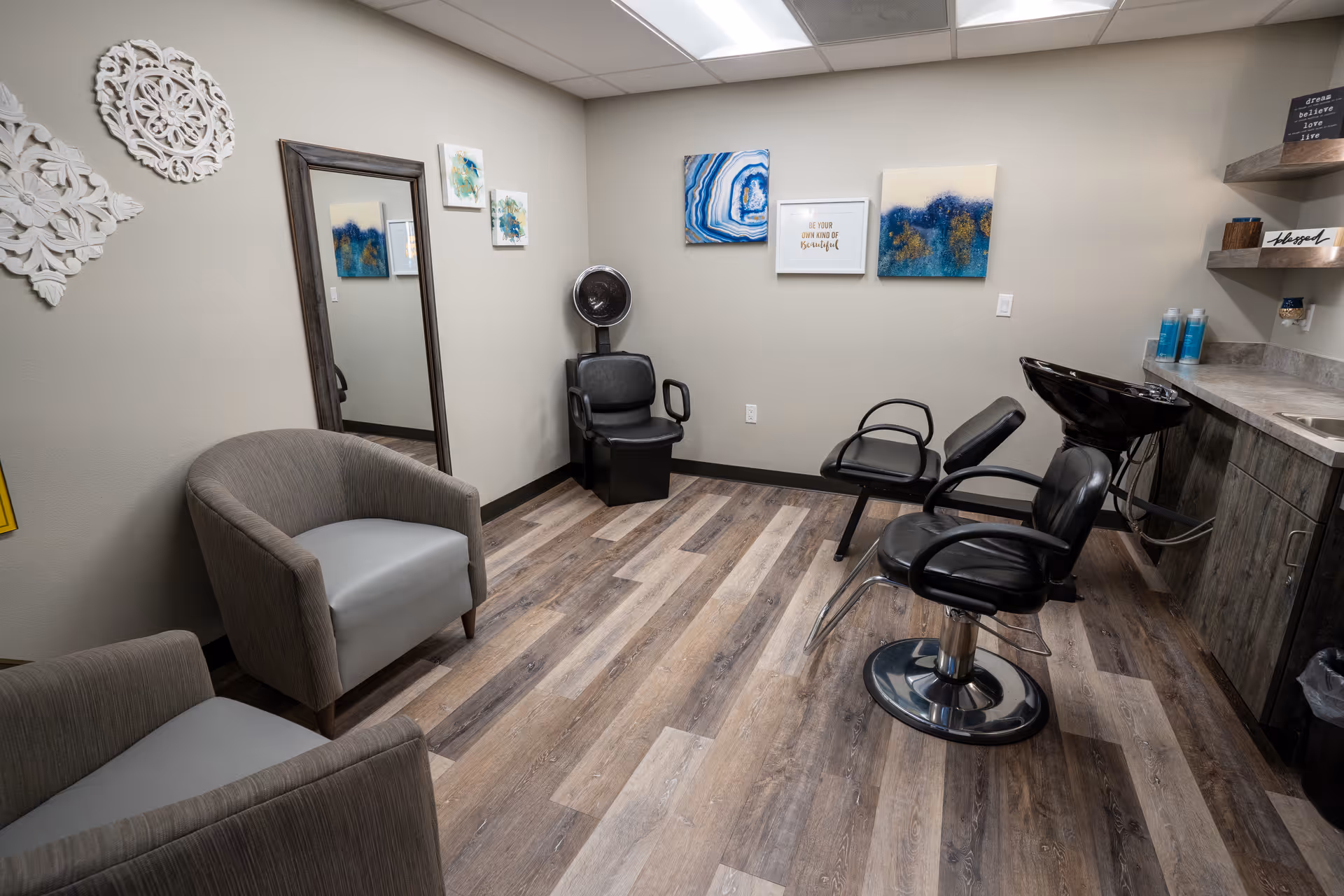 Interior view of a small salon room with two salon chairs, a hair washing station, a black hair dryer chair, two gray armchairs, a large mirror, and decorative wall art on light-colored walls with wood-patterned flooring.