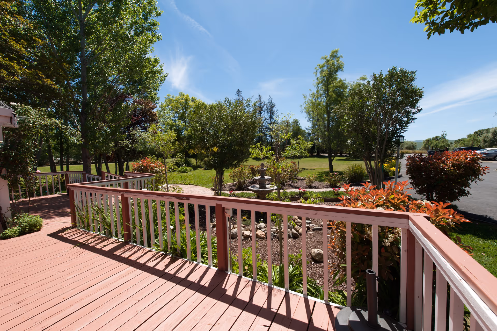 View from a wooden deck with white railings overlooking a landscaped garden with various trees, shrubs, and a small fountain under a clear blue sky.