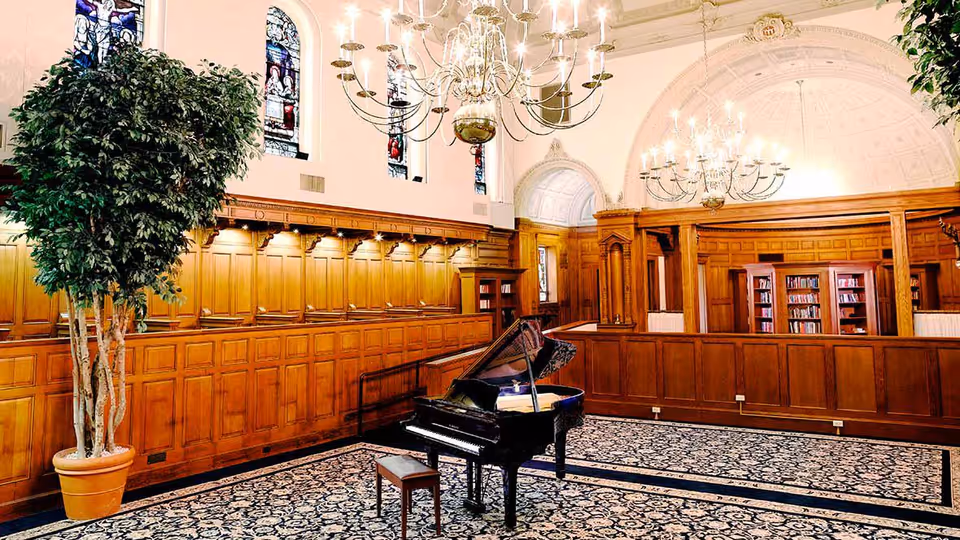 Spacious wood-paneled communal room with a grand piano on a patterned rug, chandeliers, stained-glass windows, bookshelves and a potted tree.