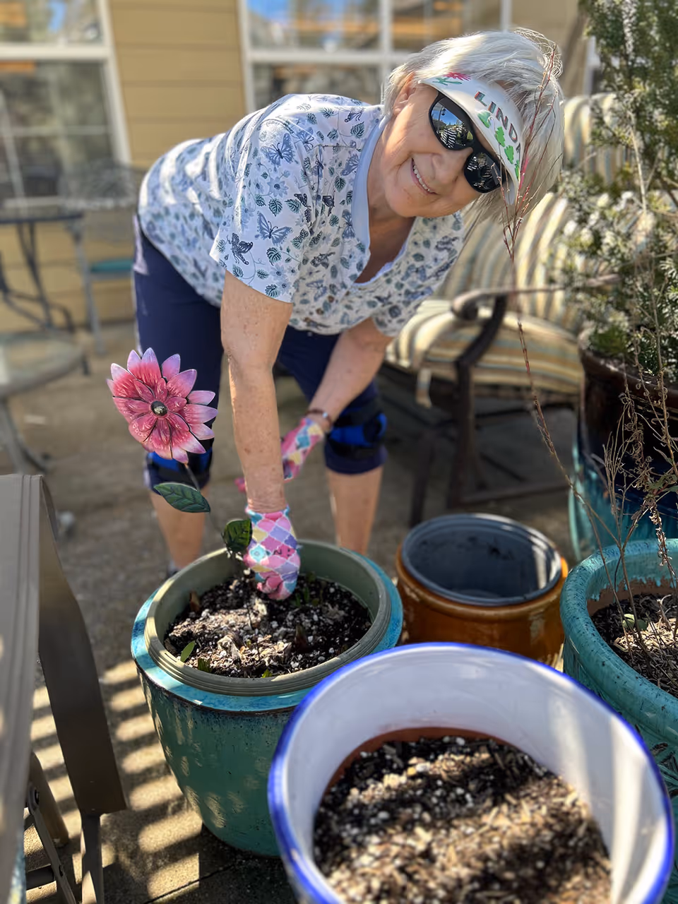 An elderly woman wearing sunglasses, a visor, and gardening gloves is tending to a potted plant outdoors on a patio. She is smiling and leaning over a large green pot filled with soil and a decorative pink flower stake. Several other empty or partially filled pots are nearby, and patio furniture is visible in the background.