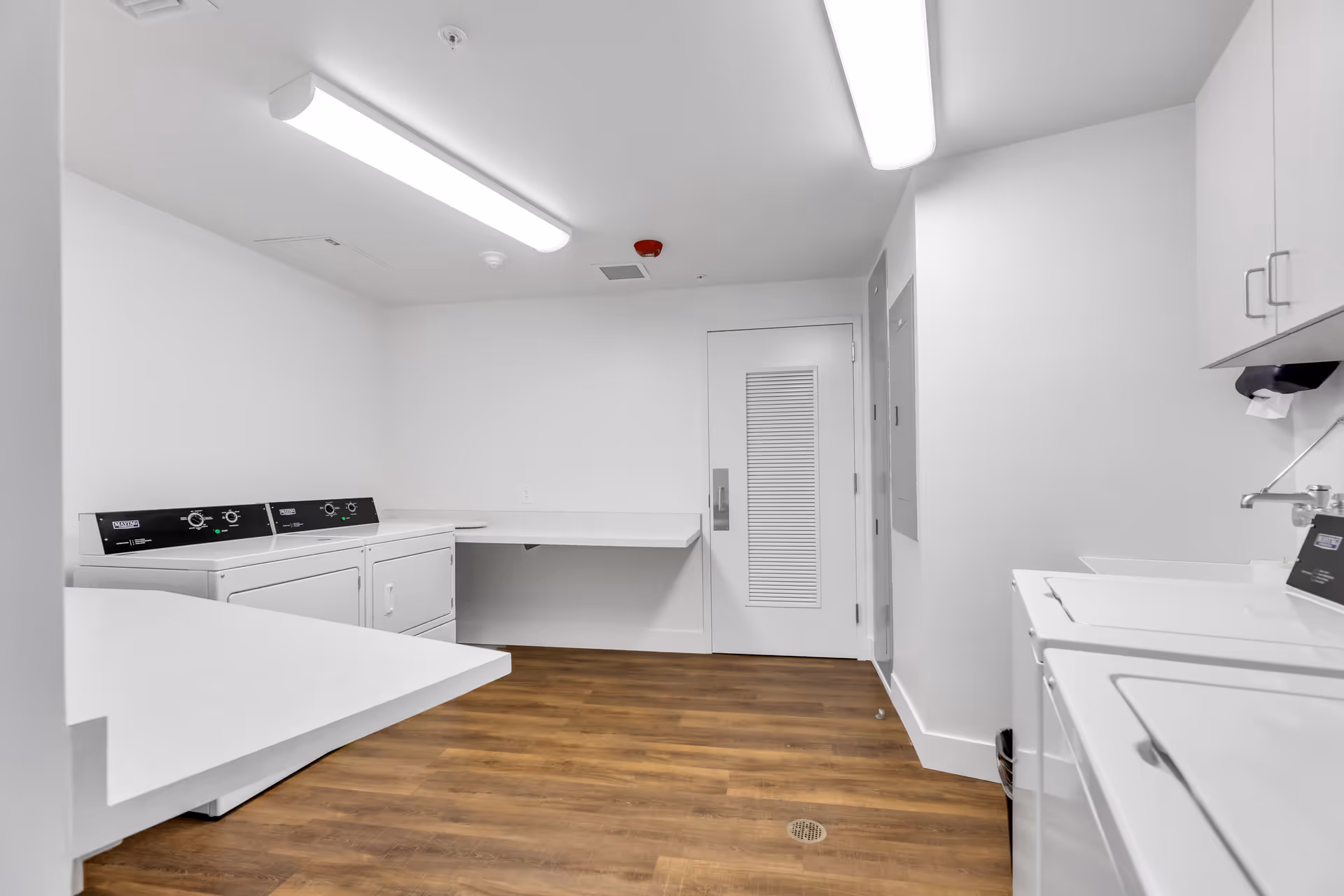 Bright, clean laundry room with washers and dryers, folding counters, white walls and wood-look flooring.