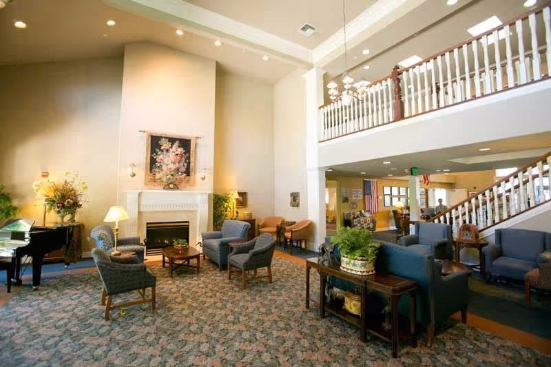 A spacious and well-lit living room area in a senior living facility featuring a high ceiling, a white fireplace with floral artwork above it, several armchairs and sofas arranged around coffee tables, a grand piano on the left, a large patterned carpet, and a staircase with white railings leading to an upper floor.