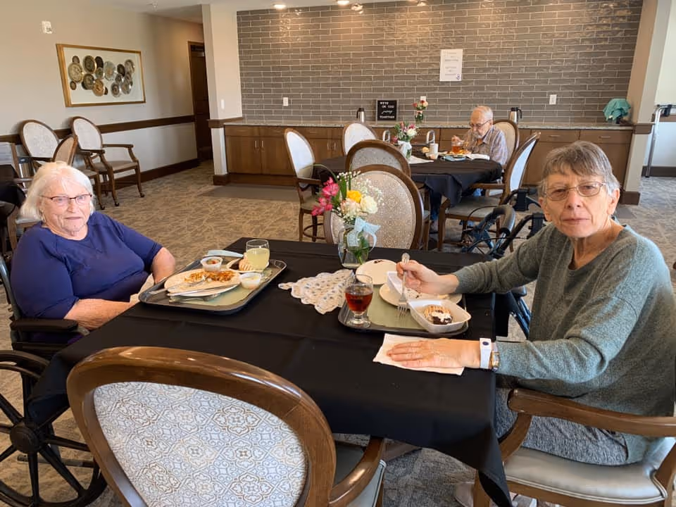 Two elderly women sitting at a dining table in a senior living facility, eating a meal. One woman is wearing a blue shirt and glasses, and the other is wearing a green sweater and glasses. There is a vase with flowers on the table, and a man is seated at another table in the background also eating. The room has carpeted floors, cushioned chairs, and a wall with gray tiles and cabinets.