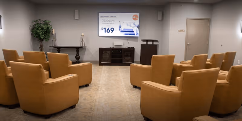 A small conference or presentation room with two rows of tan leather chairs facing a wall-mounted TV screen displaying a mattress advertisement. The room has neutral-colored walls, a carpeted floor, a potted plant, a small table with candles, and a podium near a closed door.