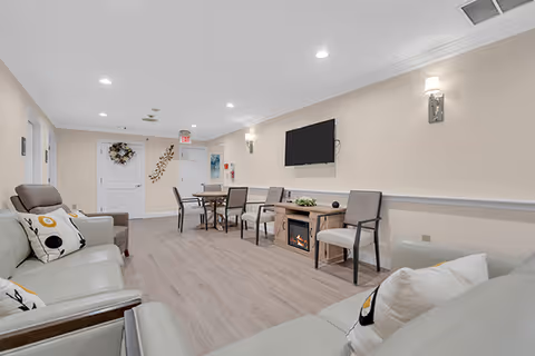 Bright communal lounge with sofas, chairs, a wall-mounted TV, an electric fireplace, and a small dining table.