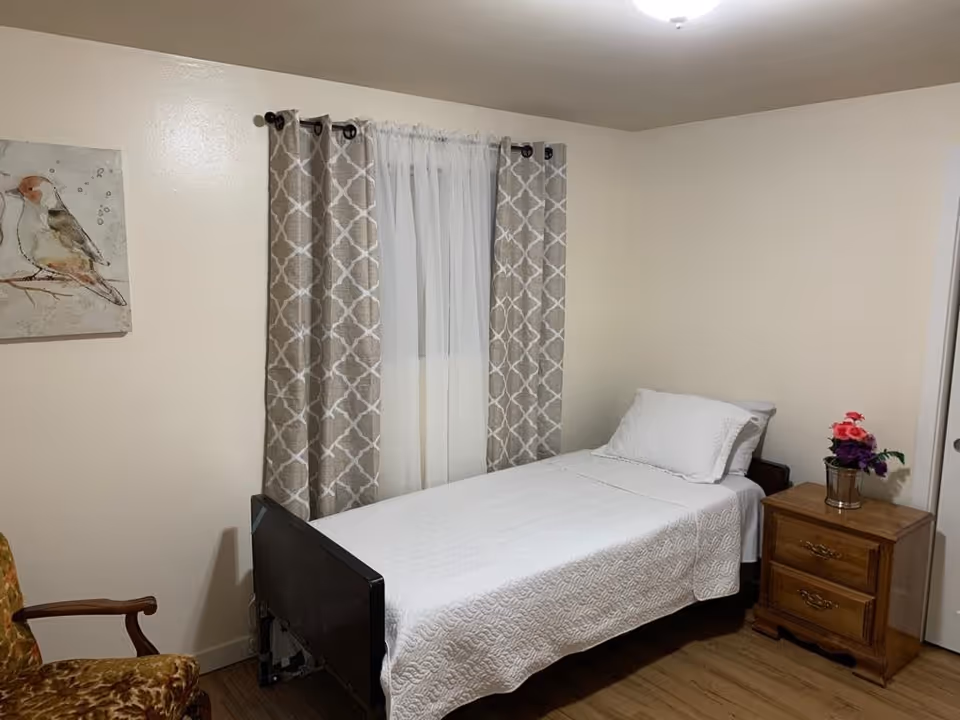 A small bedroom with a single bed covered in a white quilt and pillow. Next to the bed is a wooden nightstand with two drawers and a small flower arrangement on top. There is a window with sheer white curtains and patterned beige drapes. A framed picture of a bird hangs on the wall, and a wooden armchair with a floral cushion is partially visible.